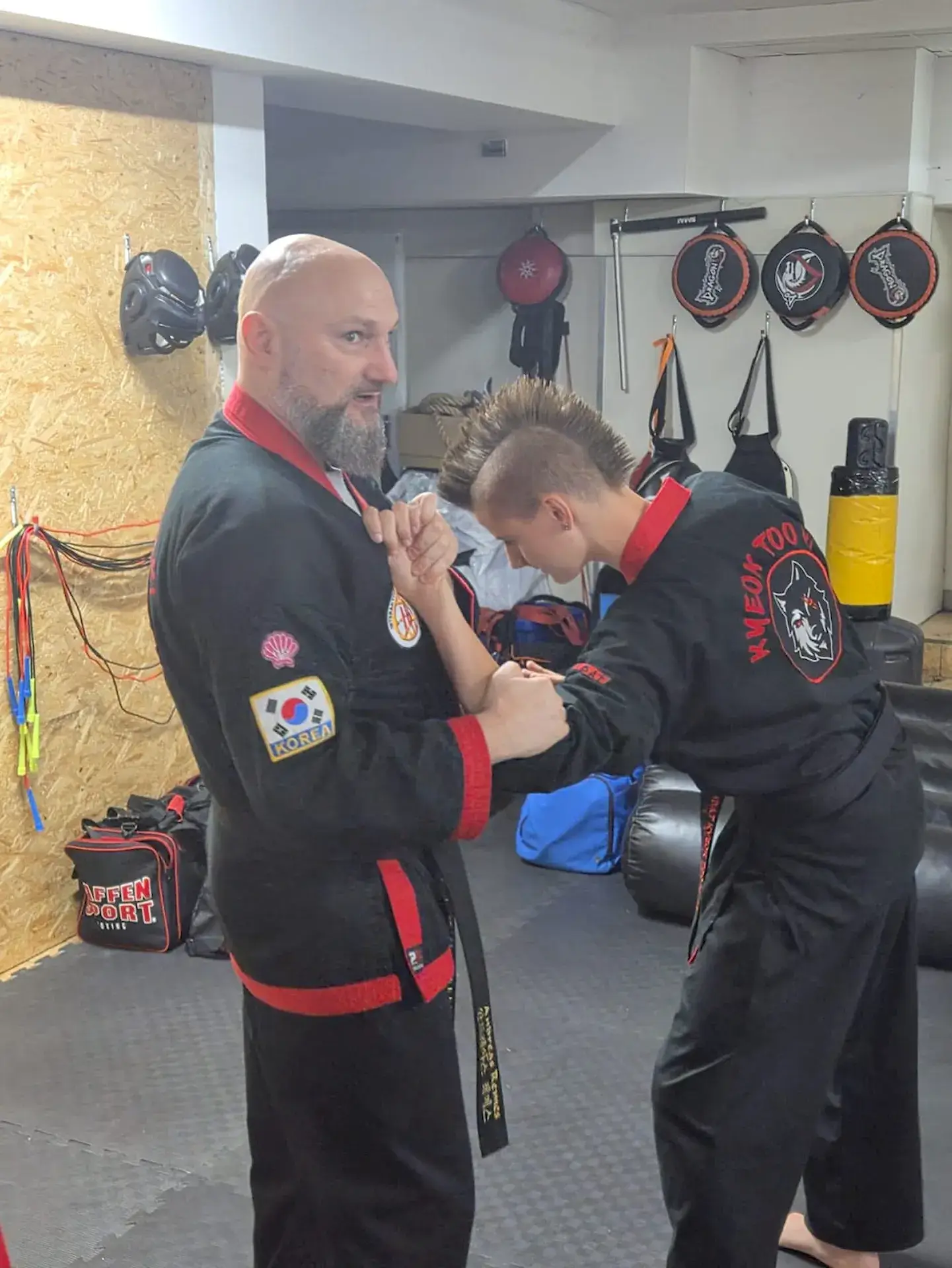 Zwei Kampfsportler in schwarzen Anzügen mit roten Besätzen demonstrieren eine Griffkontrolle am Arm.