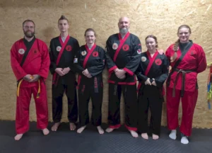 Sechs Kampfsportlerinnen und Kampfsportler in schwarzen und roten Anzügen stehen für ein Gruppenfoto nebeneinander.