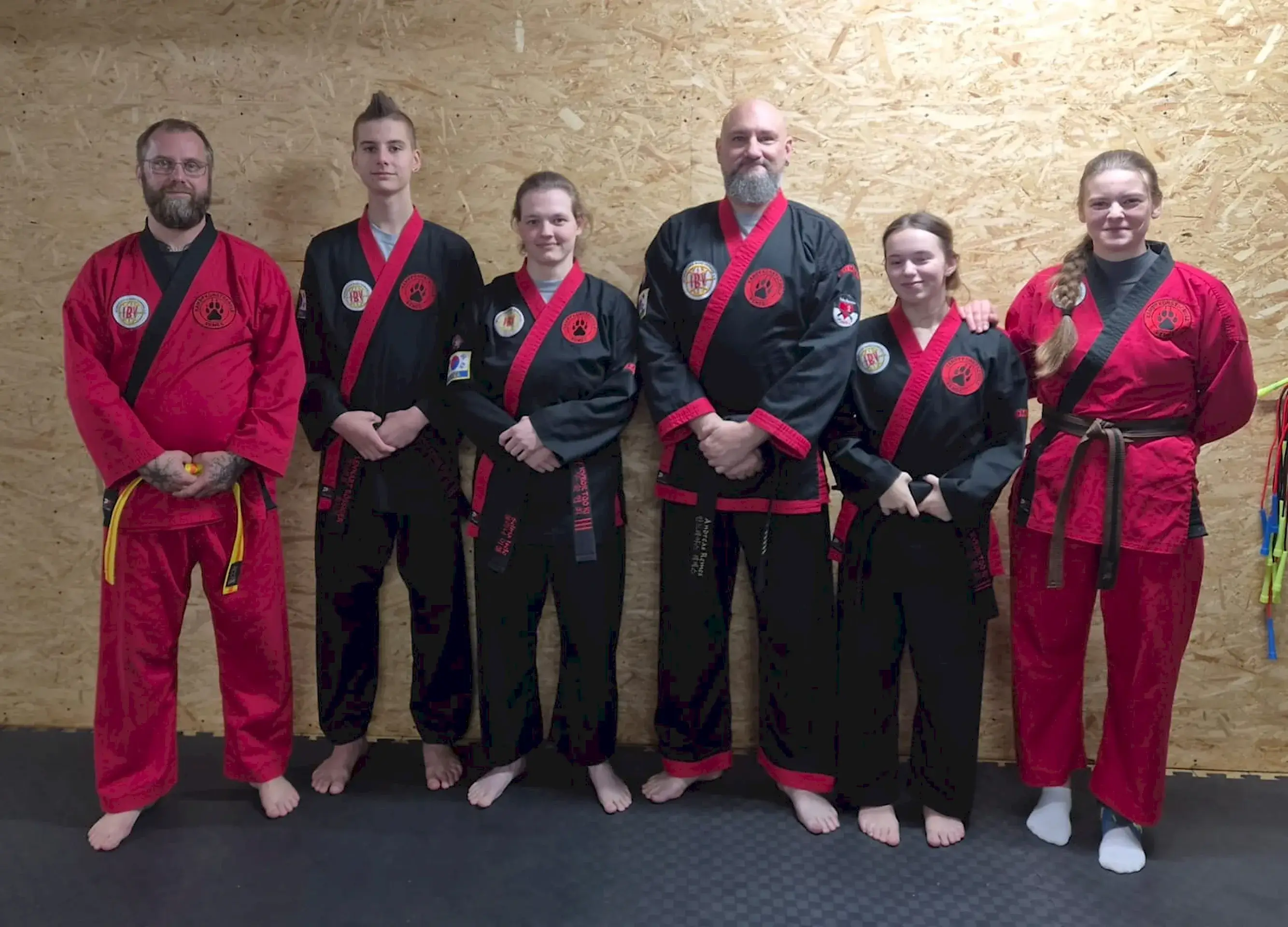 Sechs Kampfsportlerinnen und Kampfsportler in schwarzen und roten Anzügen stehen für ein Gruppenfoto nebeneinander.